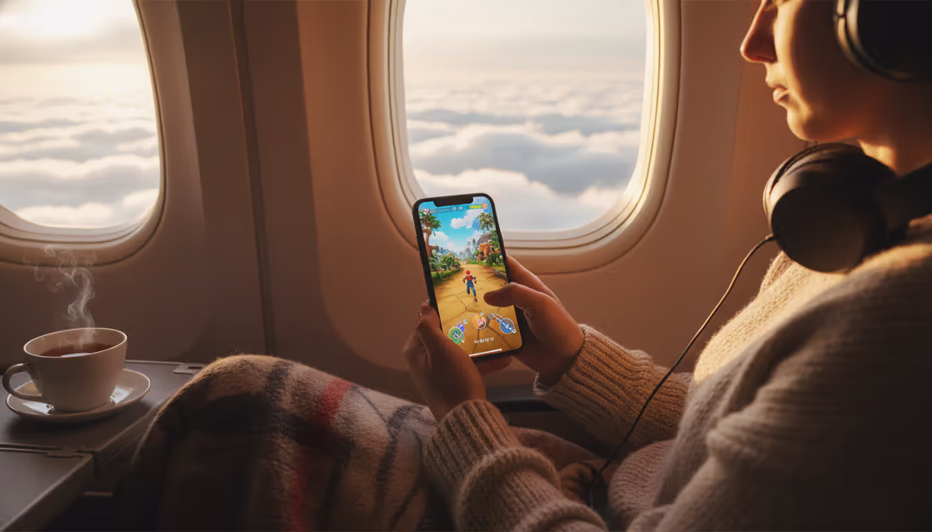A person sitting in an airplane window seat playing a game on an iPhone with clouds and sunlight visible through the window creating a warm cozy travel atmosphere