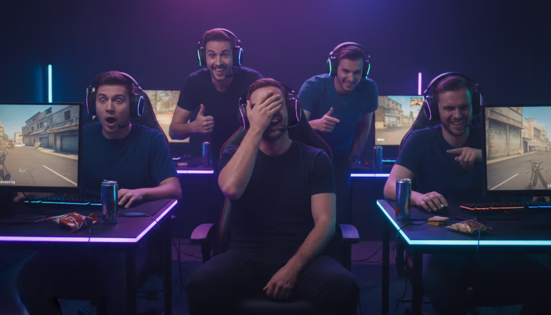 Five gamers sitting at desks with glowing monitors in a dark neon-lit room, laughing and reacting emotionally during a first-person shooter match